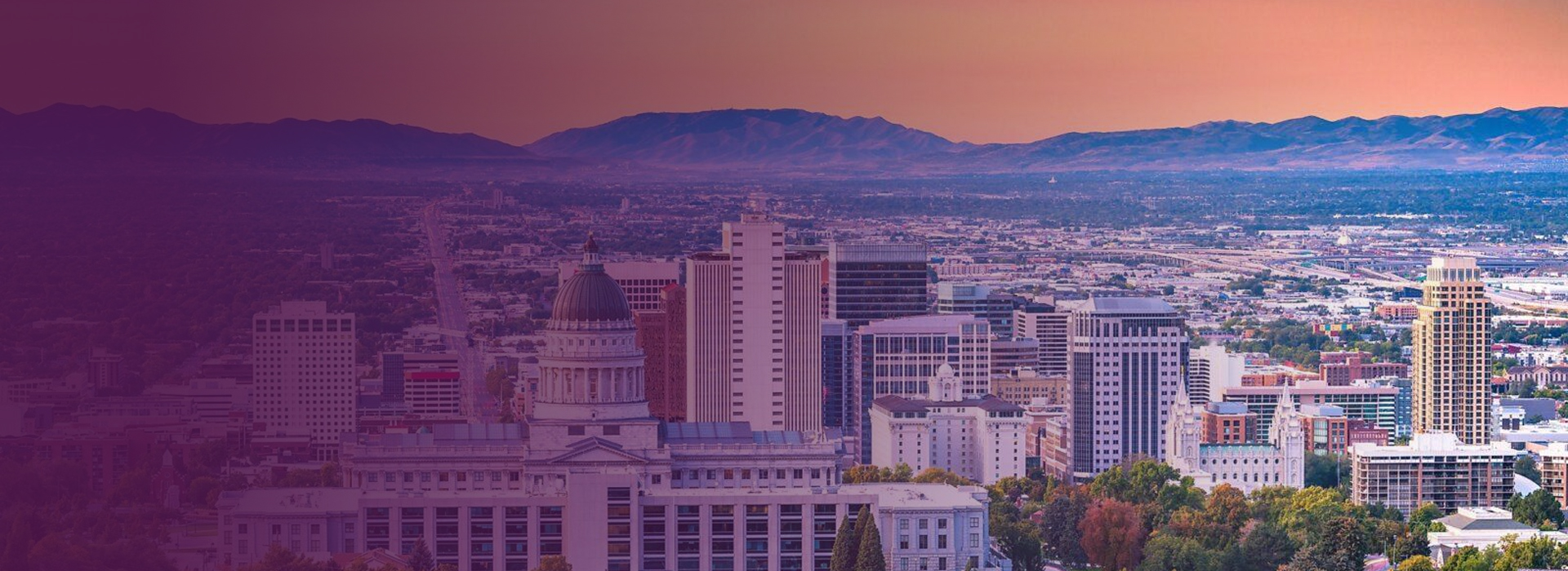 City skyline with Utah State Capitol building in foreground and mountains in the background at sunset.