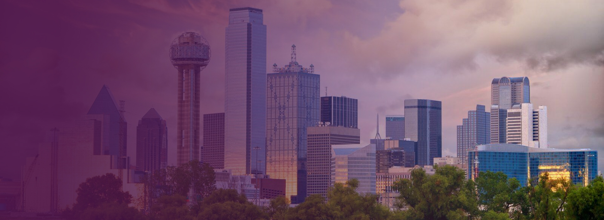 Dallas city skyline at sunset with tall modern buildings and green trees in the foreground.