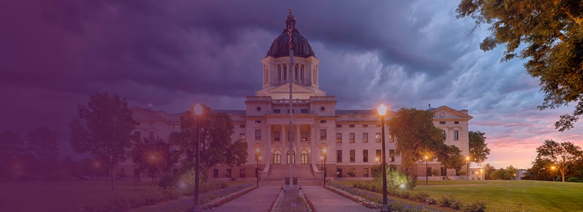 South Dakota State Capitol building under a dramatic evening sky with illuminated street lamps and green lawns.