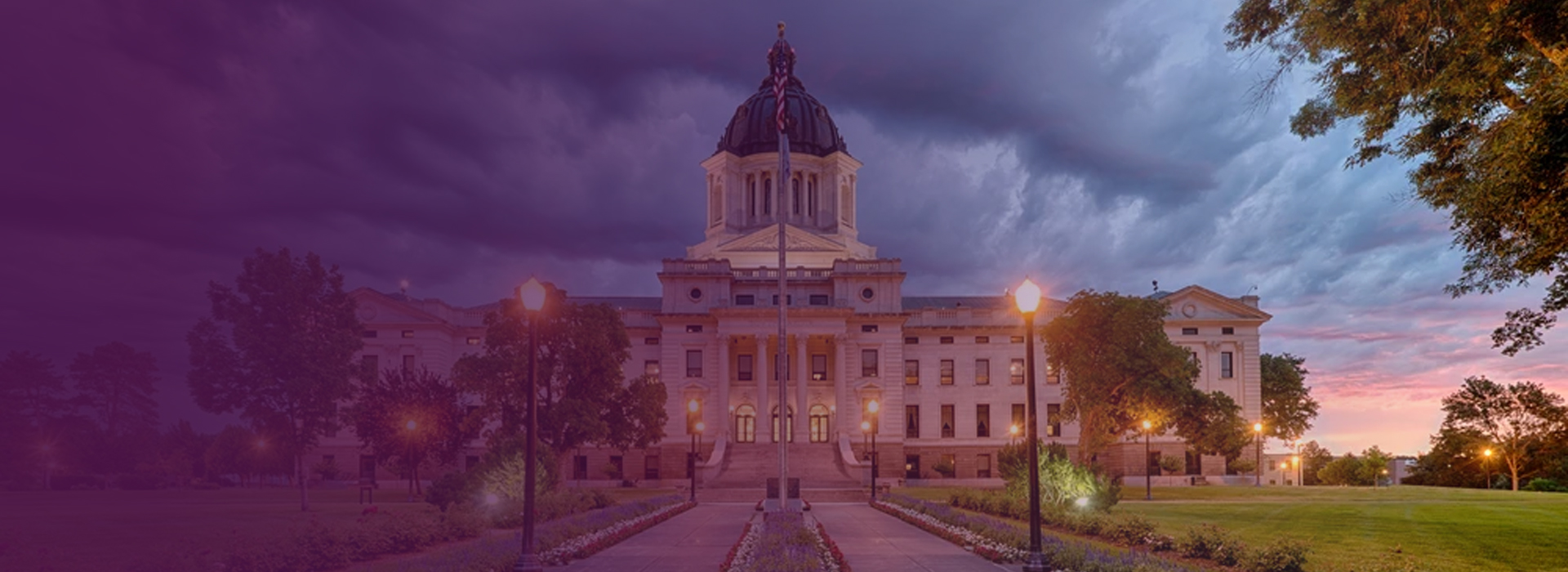 South Dakota State Capitol building under a dramatic evening sky with illuminated street lamps and green lawns.
