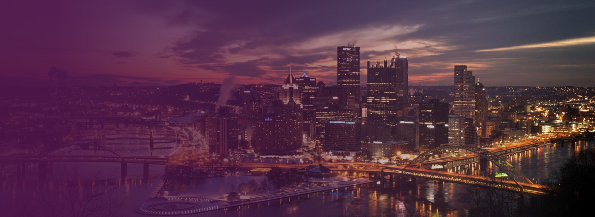 Nighttime cityscape of Pittsburgh, Pennsylvania, with illuminated buildings and bridges over a river under a cloudy sky.