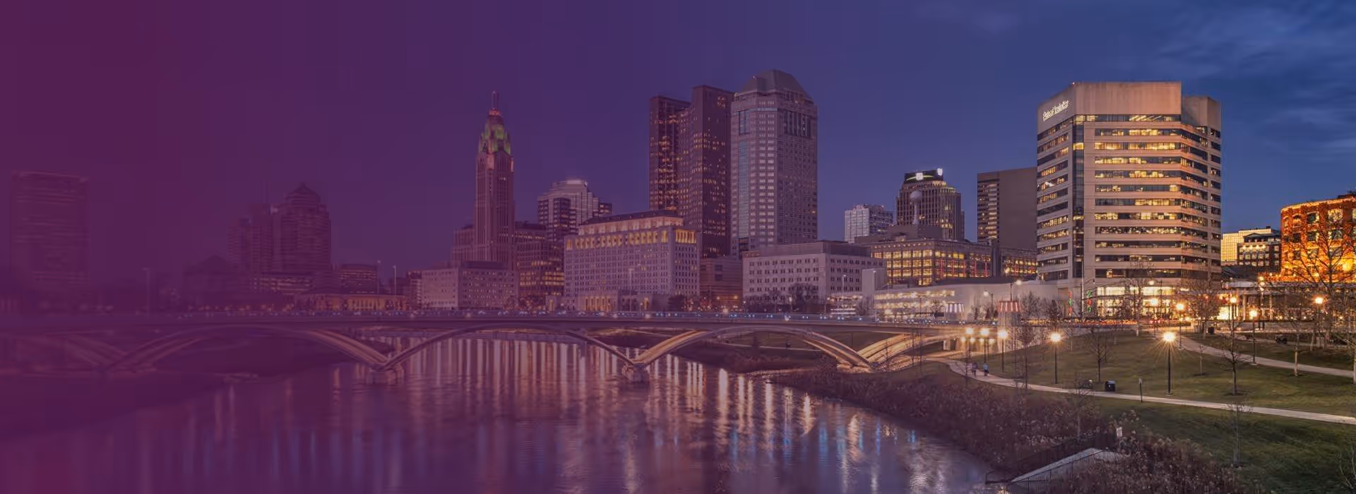 Twilight view of Columbus, Ohio skyline with illuminated buildings and a bridge over the Scioto River.