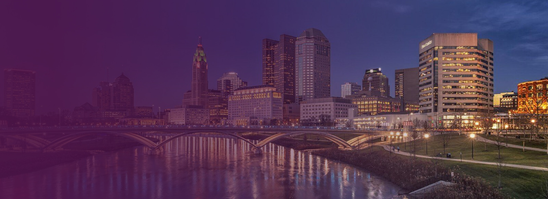 Twilight view of Columbus, Ohio skyline with illuminated buildings and a bridge over the Scioto River.