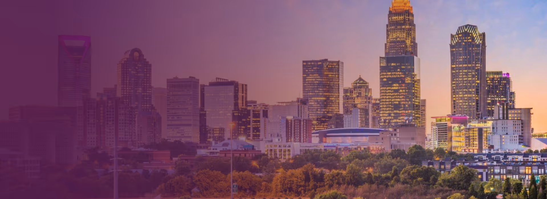 Sunset view of Charlotte, North Carolina skyline with illuminated skyscrapers and trees in the foreground.