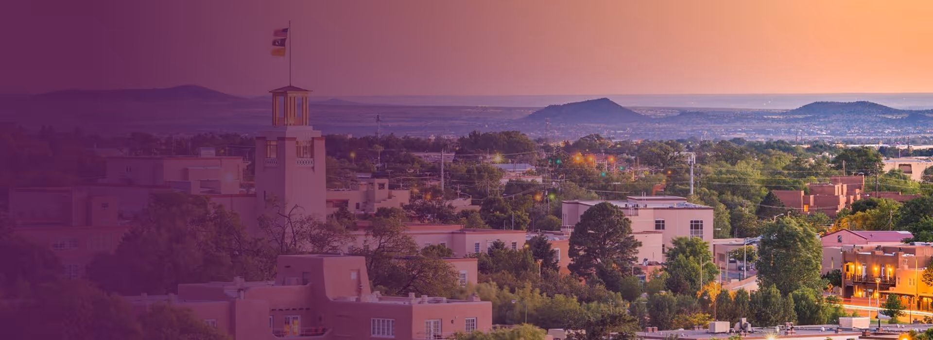 Cityscape of Santa Fe, New Mexico, featuring adobe-style buildings and distant hills at sunset.