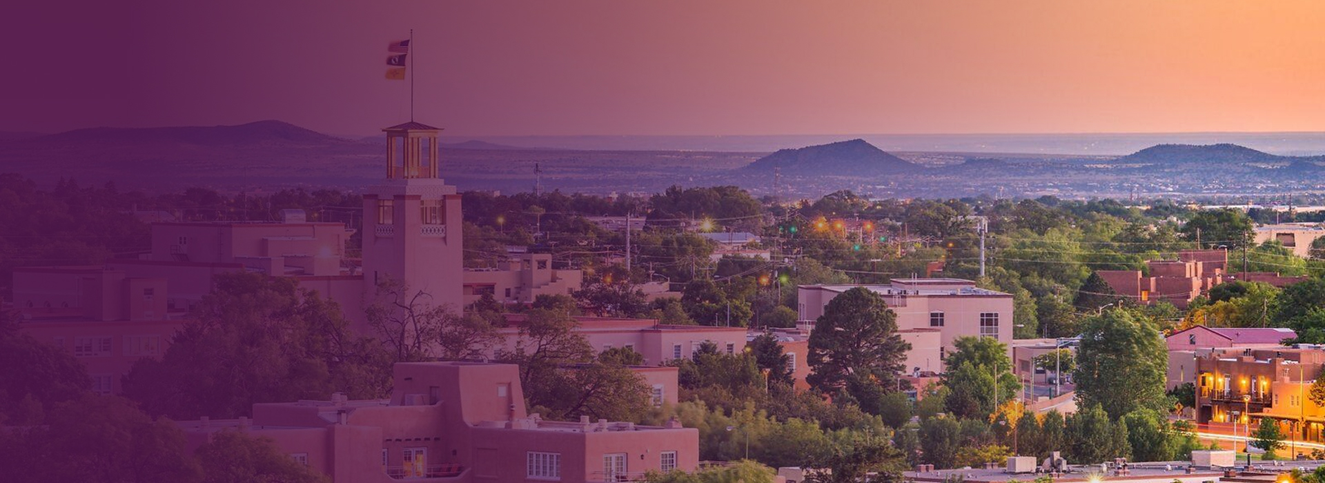 Cityscape of Santa Fe, New Mexico, featuring adobe-style buildings and distant hills at sunset.