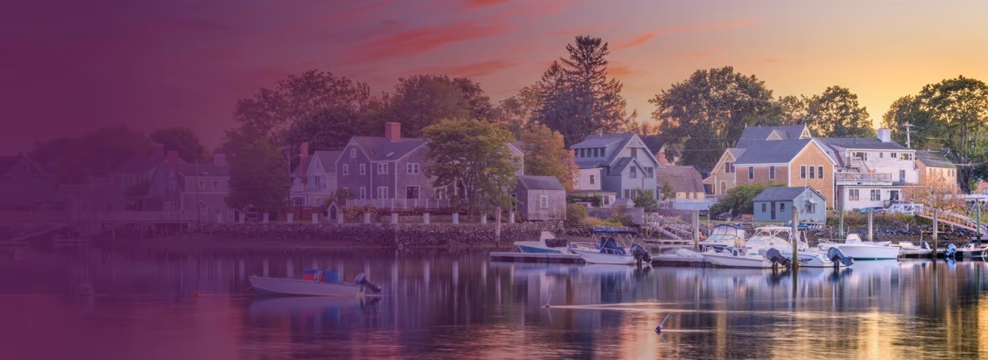 Calm harbor at sunset with boats docked near a shoreline village with trees and quaint houses.