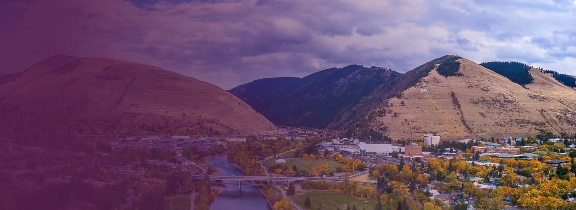 Mountains with an 'M' on one slope overlooking a river, bridges, and a town with autumn trees.