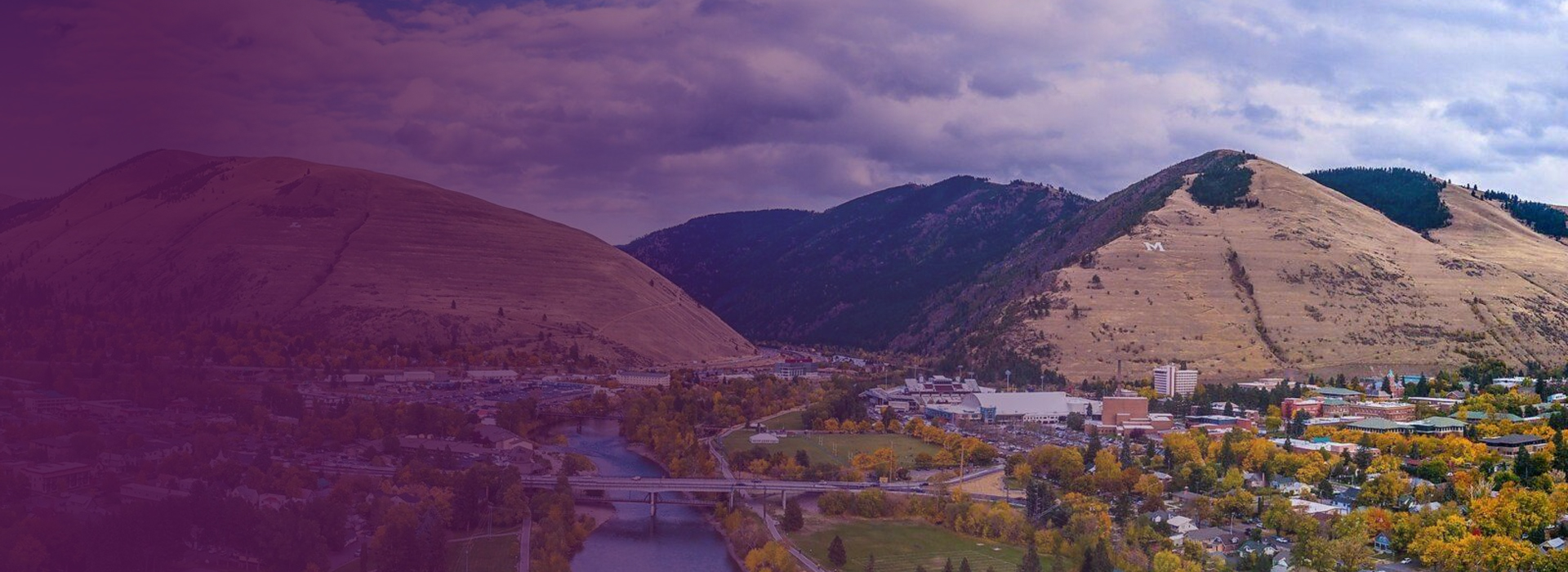 Mountains with an 'M' on one slope overlooking a river, bridges, and a town with autumn trees.