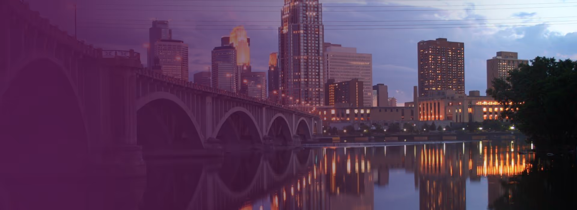 City skyline at dusk with a multi-arched bridge over calm river reflecting building lights.