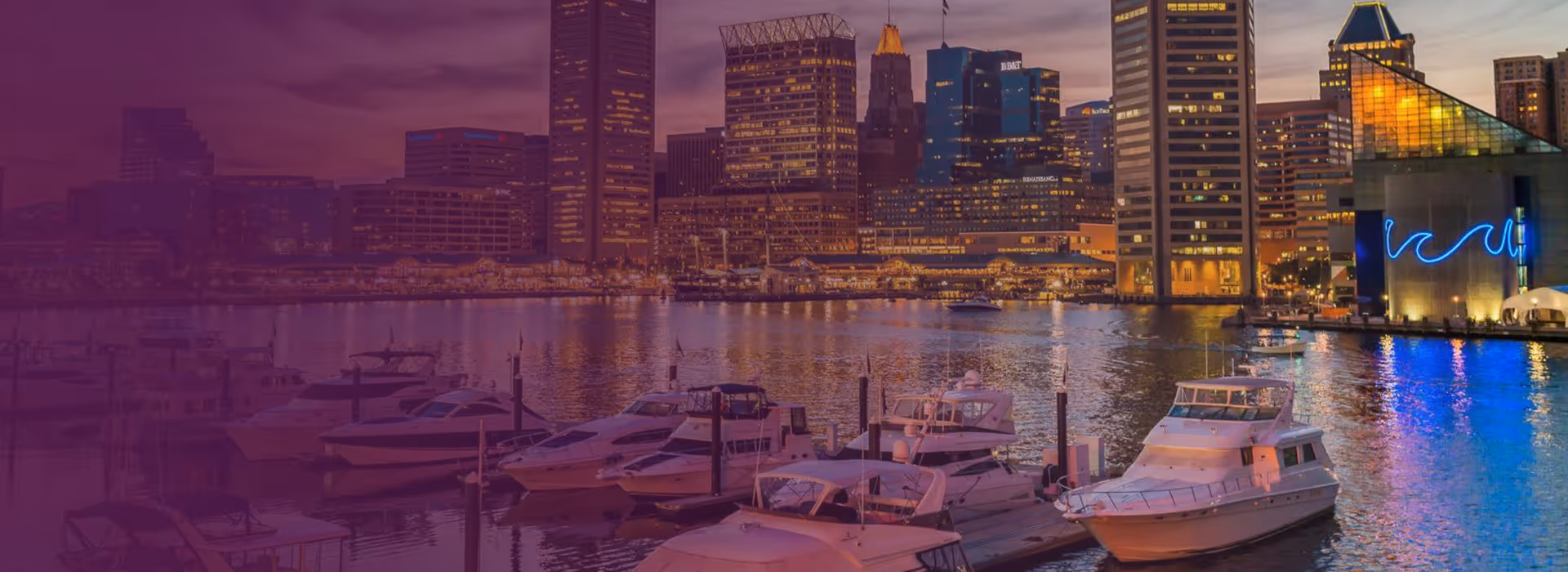 Marina with docked boats in front of a city skyline at dusk with illuminated buildings and reflections on the water.