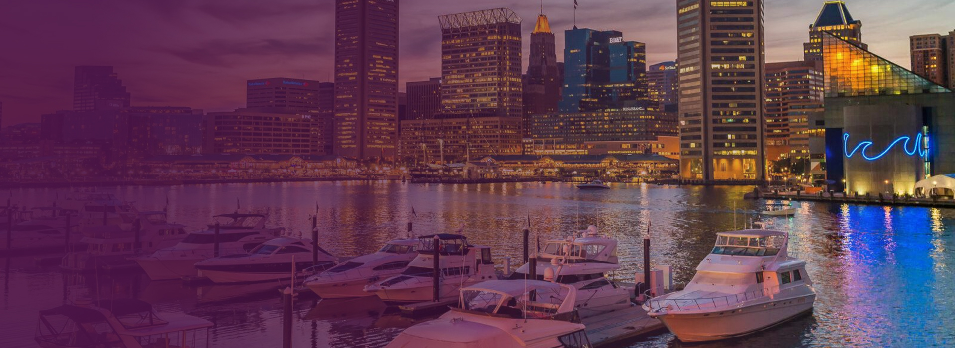 Marina with docked boats in front of a city skyline at dusk with illuminated buildings and reflections on the water.