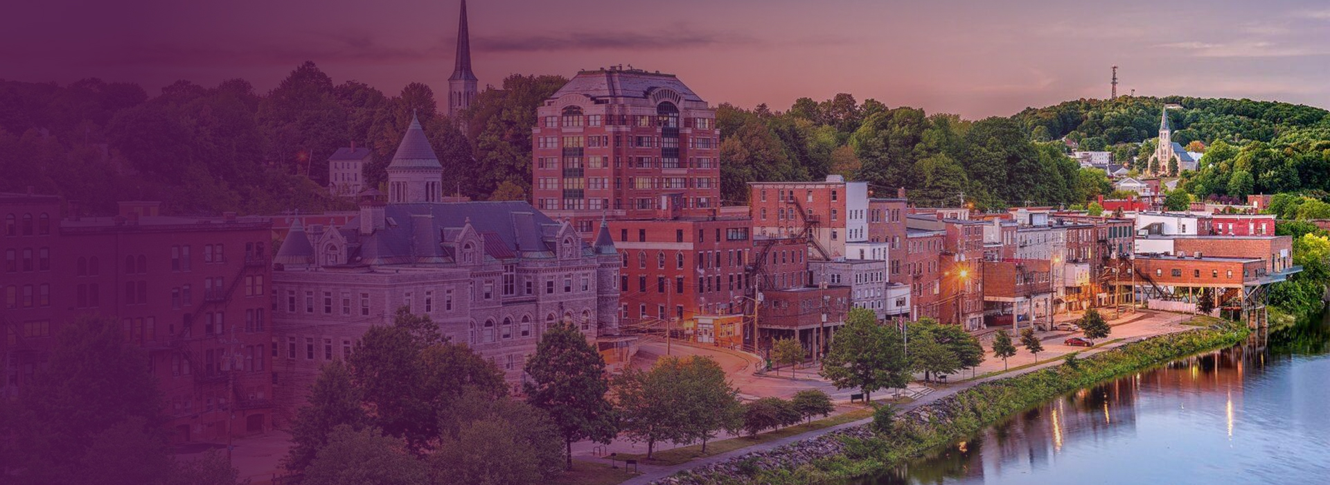 Panoramic view of a riverside town with historic buildings, trees, and a church steeple at sunset.