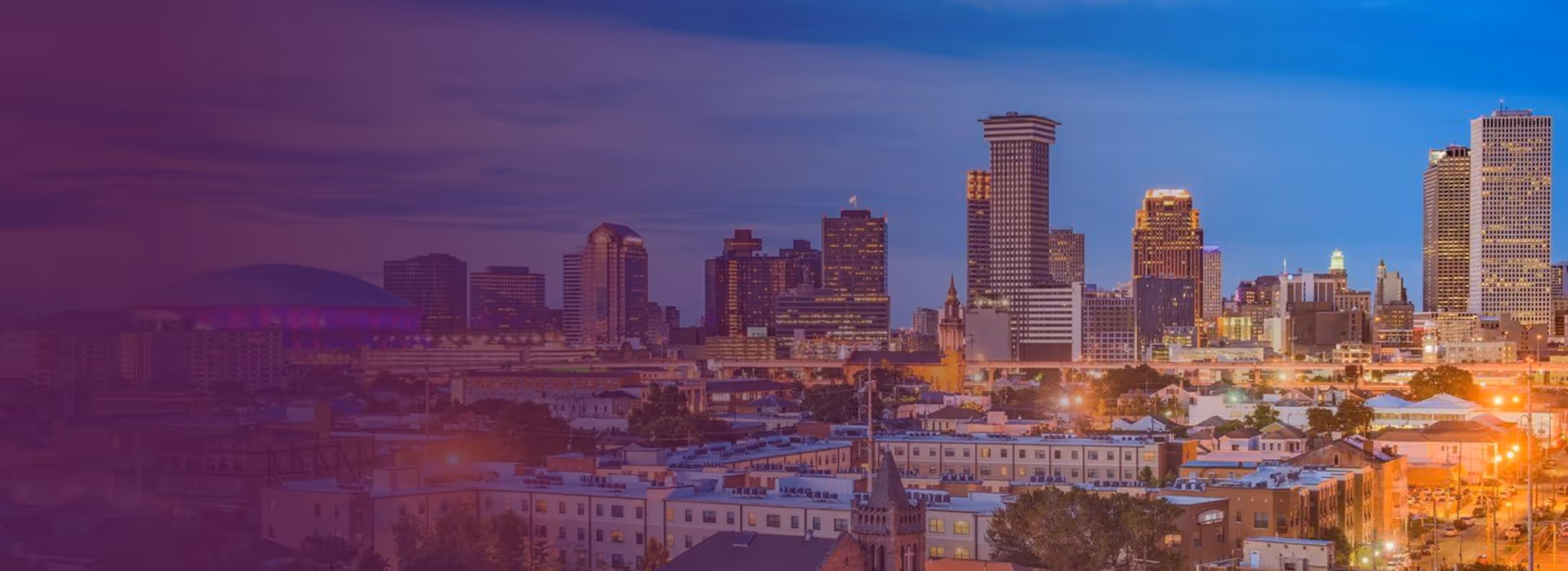 Panoramic view of New Orleans skyline at dusk with illuminated buildings and a purple-lit stadium dome.