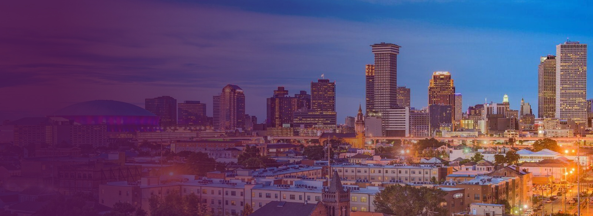Panoramic view of New Orleans skyline at dusk with illuminated buildings and a purple-lit stadium dome.