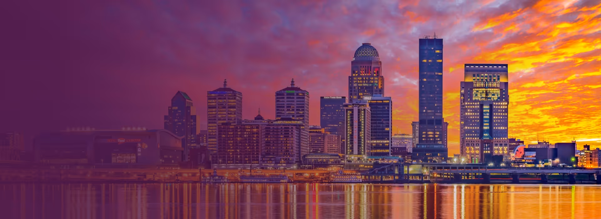 City skyline of Louisville, Kentucky at sunset with illuminated buildings reflecting on the river.