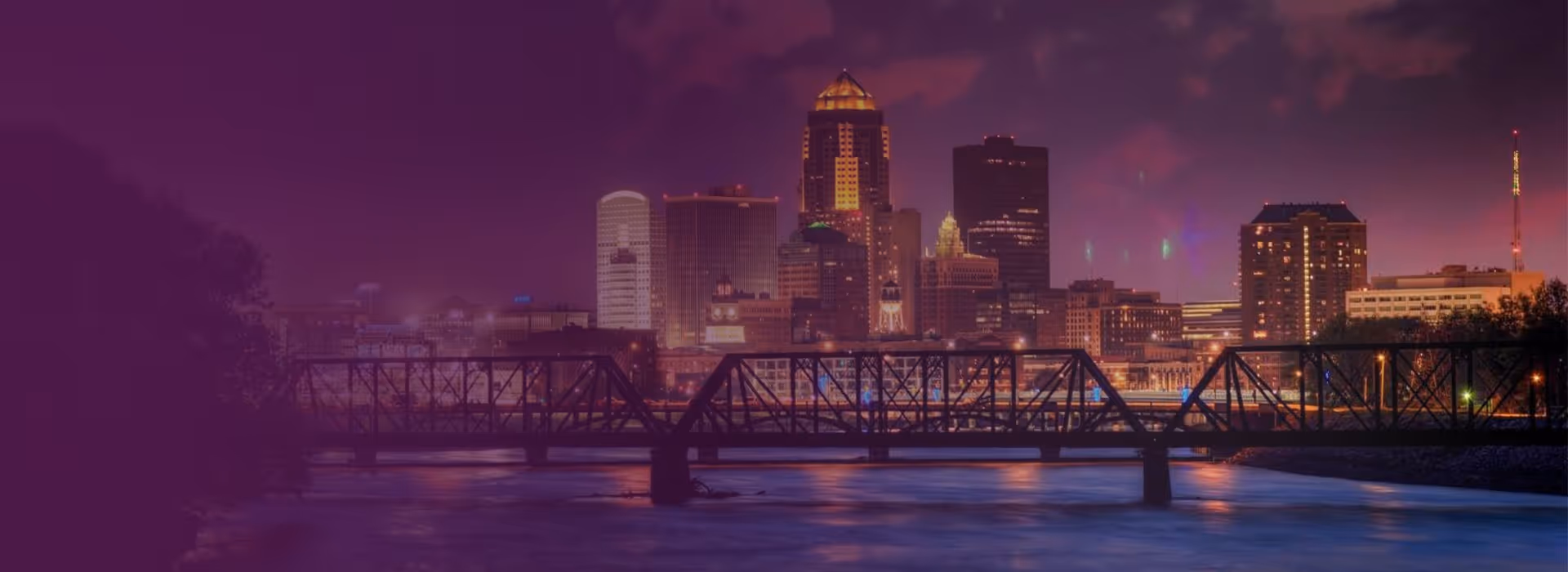 Nighttime city skyline with illuminated buildings and a bridge over a river in the foreground.