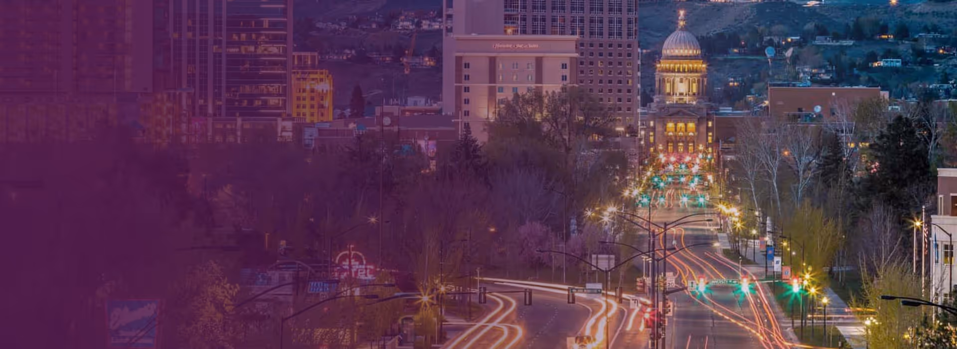 City street at dusk with light trails from cars and a domed capitol building in the background, surrounded by trees and tall buildings.