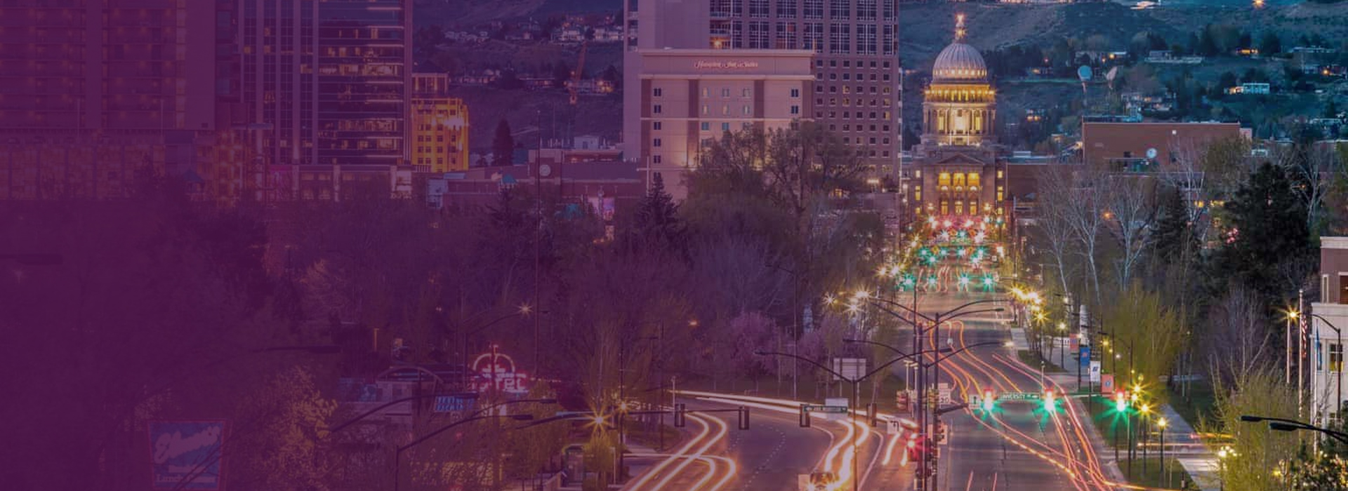 City street at dusk with light trails from cars and a domed capitol building in the background, surrounded by trees and tall buildings.