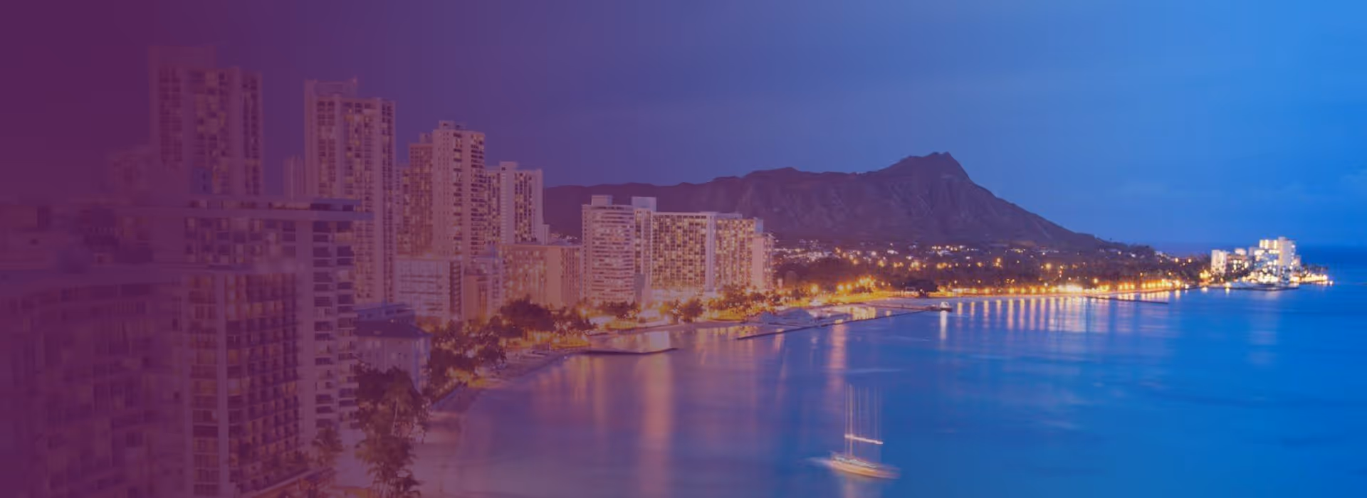 Evening view of Waikiki skyline with lit buildings along the beach and Diamond Head crater in the background in Honolulu, Hawaii.