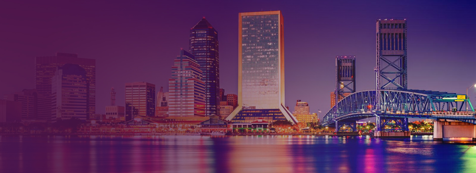 Night skyline of Jacksonville, Florida, with illuminated buildings and a lit steel bridge over calm water reflecting city lights.