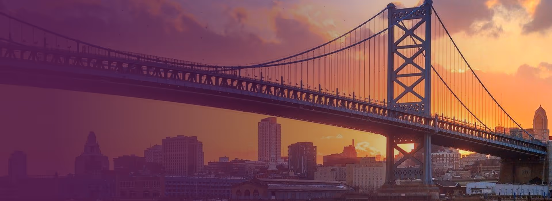Suspension bridge over a cityscape at sunset with an orange sky.