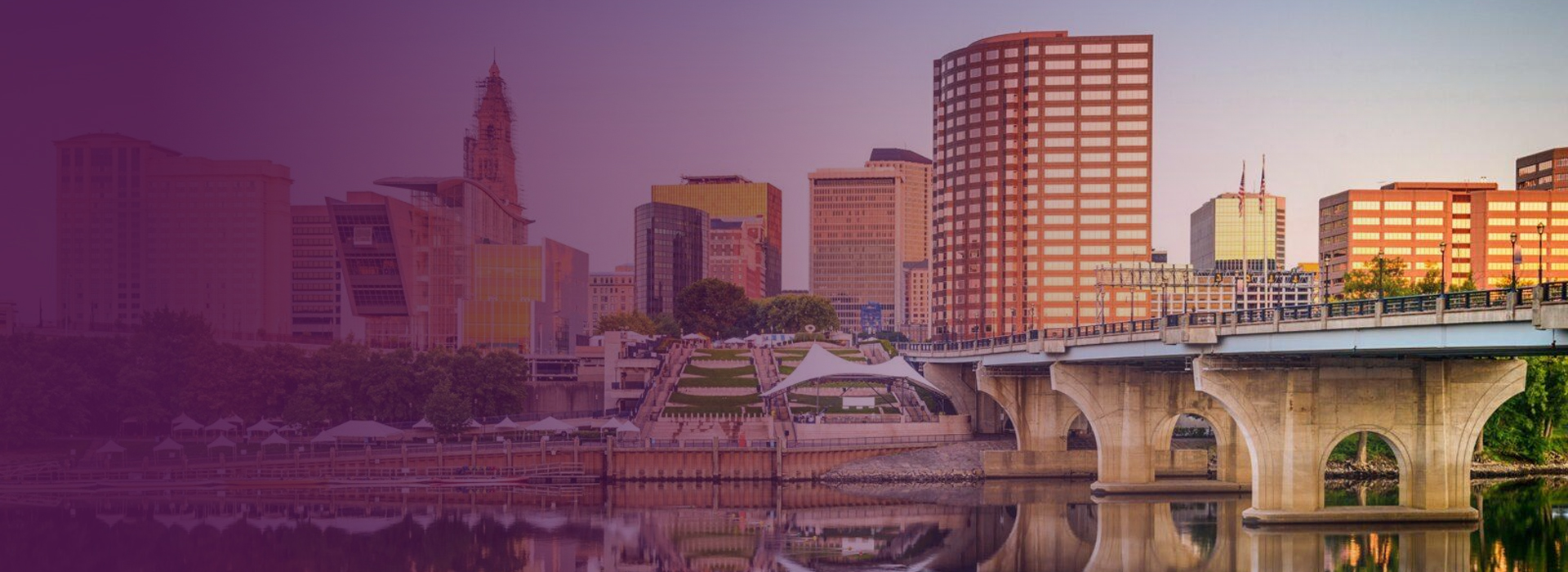 City skyline with modern buildings and a concrete bridge reflected in calm river water at sunset.