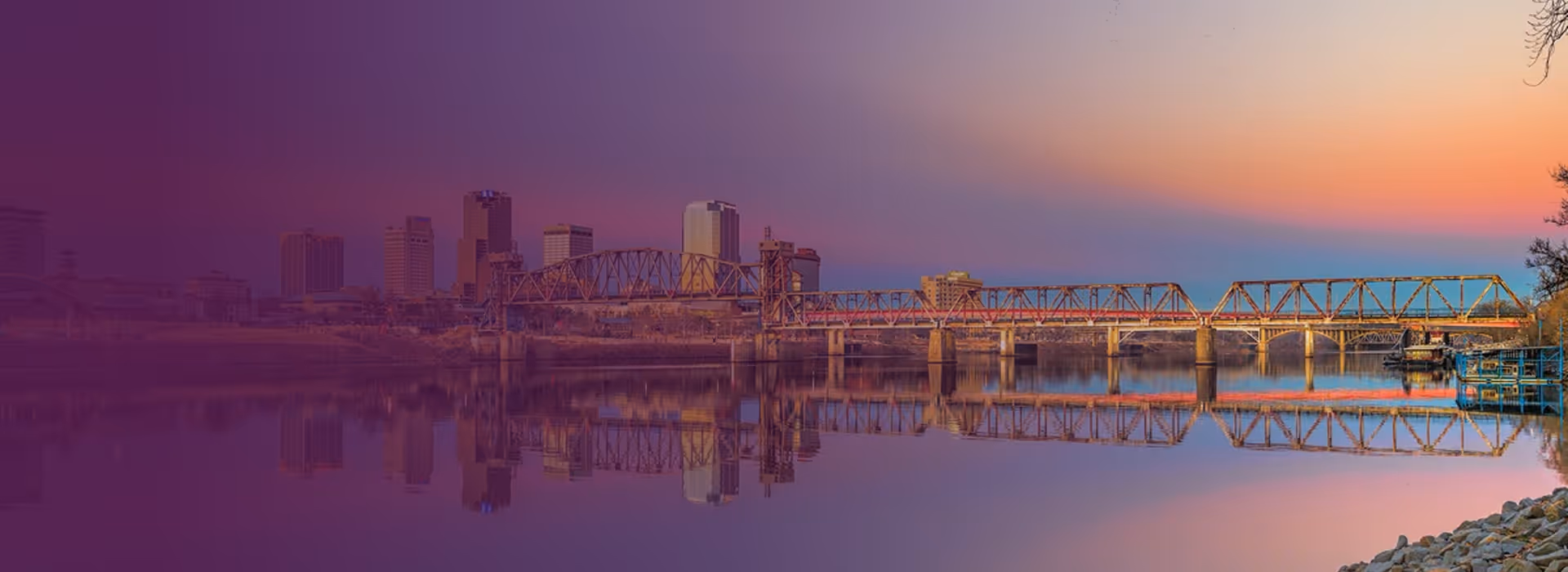City skyline with a truss bridge over a calm river at sunset, reflecting buildings and the sky.