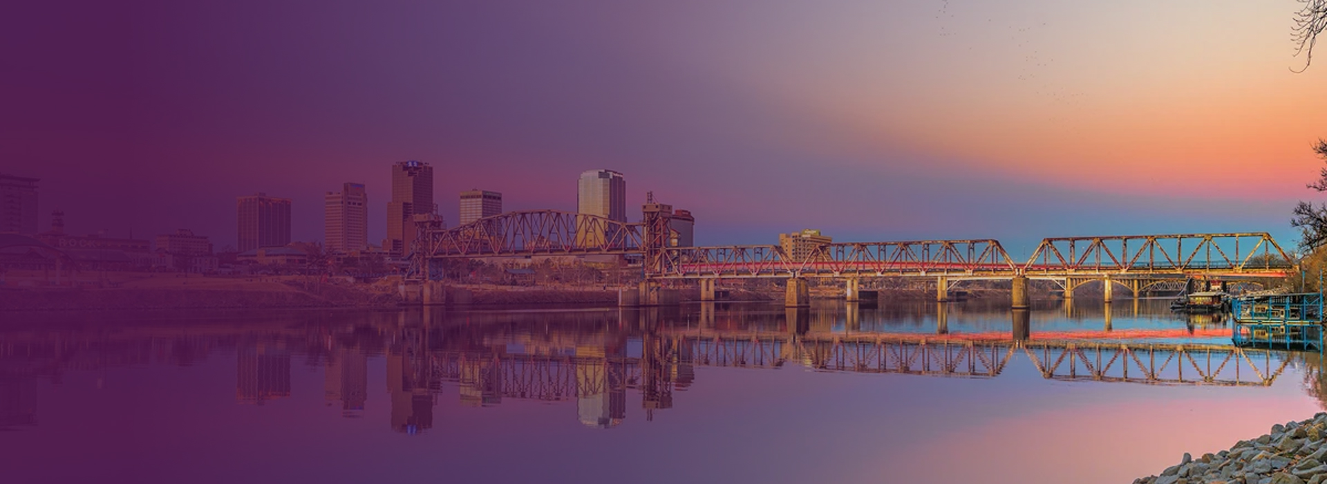 City skyline with a truss bridge over a calm river at sunset, reflecting buildings and the sky.