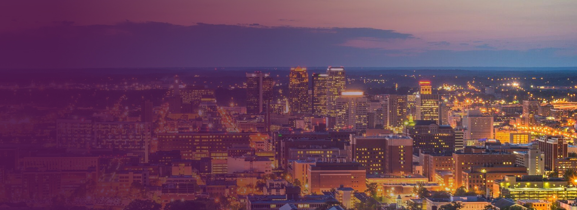 Panoramic view of a city skyline at dusk with illuminated buildings and a partly cloudy sky.