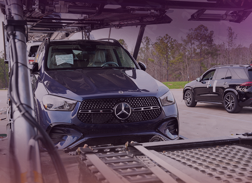 Blue Mercedes-Benz SUV loaded onto an open car transport trailer with a black SUV parked nearby in an outdoor lot.