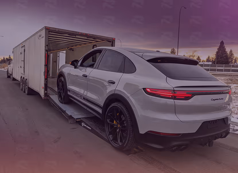 White Porsche Cayenne Turbo SUV being loaded into a large enclosed car trailer on a road at dusk.