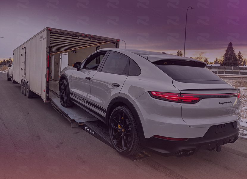 White Porsche Cayenne Turbo SUV being loaded into a large enclosed car trailer on a road at dusk.