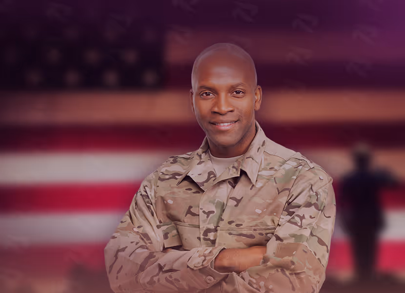 Smiling African American soldier in camouflage uniform standing with arms crossed in front of a blurred American flag background.