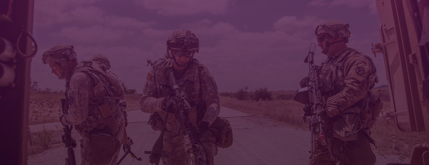 Four soldiers in full combat gear standing on a dirt road holding rifles, in a rural outdoor setting.
