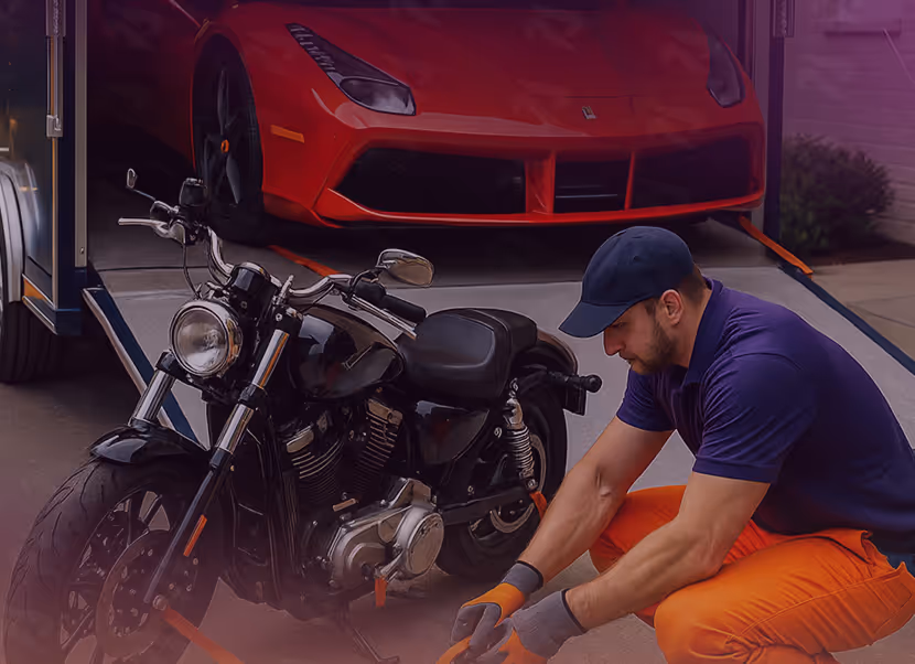 Man wearing a navy cap, purple shirt, and orange pants securing a black motorcycle with straps in front of a red sports car loaded on a trailer.