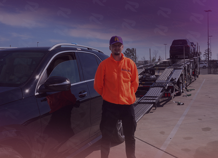 Man in an orange jacket and purple cap standing beside a black SUV and a vehicle transport trailer in a parking lot.