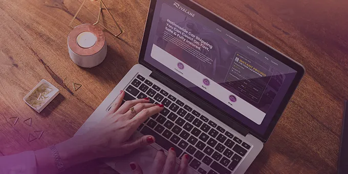 Person typing on a laptop displaying a car shipping price calculator webpage on a wooden desk with a candle and paper clips nearby.