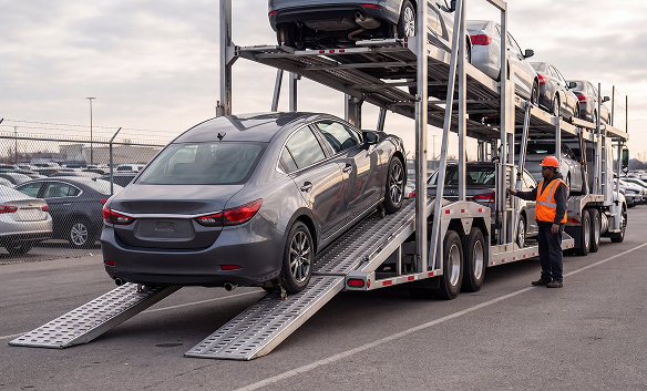 Worker in a safety vest guiding a sedan onto a car carrier trailer for auction transport.