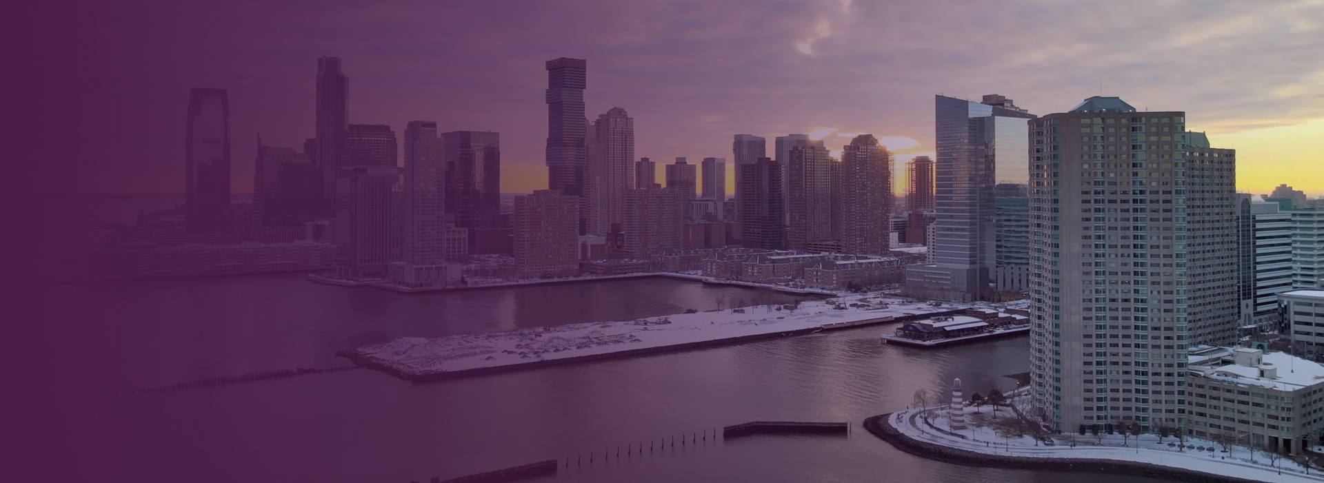 Waterfront high-rise towers at sunset seen across the water, with a pier and snow-covered shoreline in the foreground.