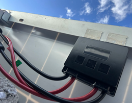 The image shows a solar panel with its wiring and control panel mounted on a metal surface under a clear blue sky.AI-generated content may be incorrect.