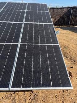 The image shows a neatly arranged solar panel array on a sandy ground, under a clear sky.AI-generated content may be incorrect.