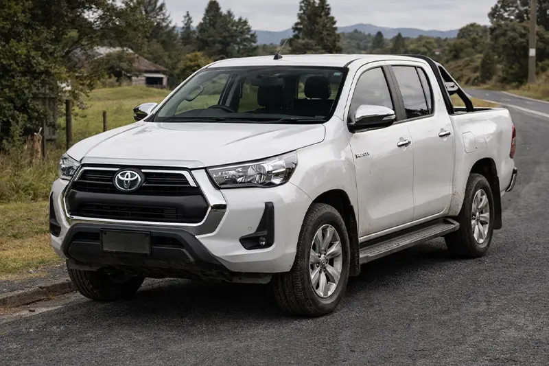 White Toyota Hilux pickup truck parked on a road near greenery and trees.