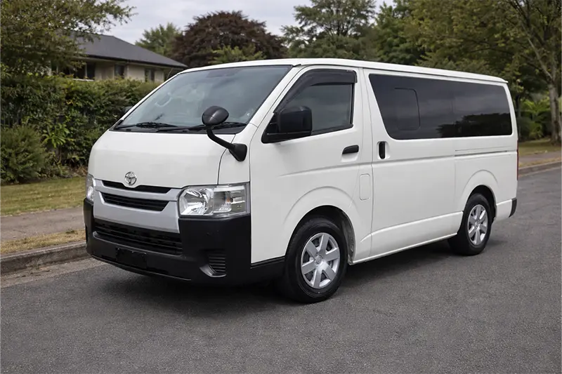 White Toyota Hiace van parked on a suburban street with greenery and houses in the background.