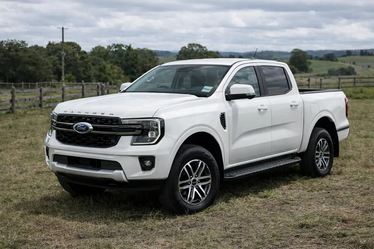 White Toyota Hilux pickup truck parked on a road near greenery and trees.