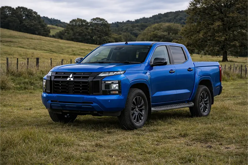 Blue Toyota Aqua compact car parked on a residential street with greenery and houses in the background.