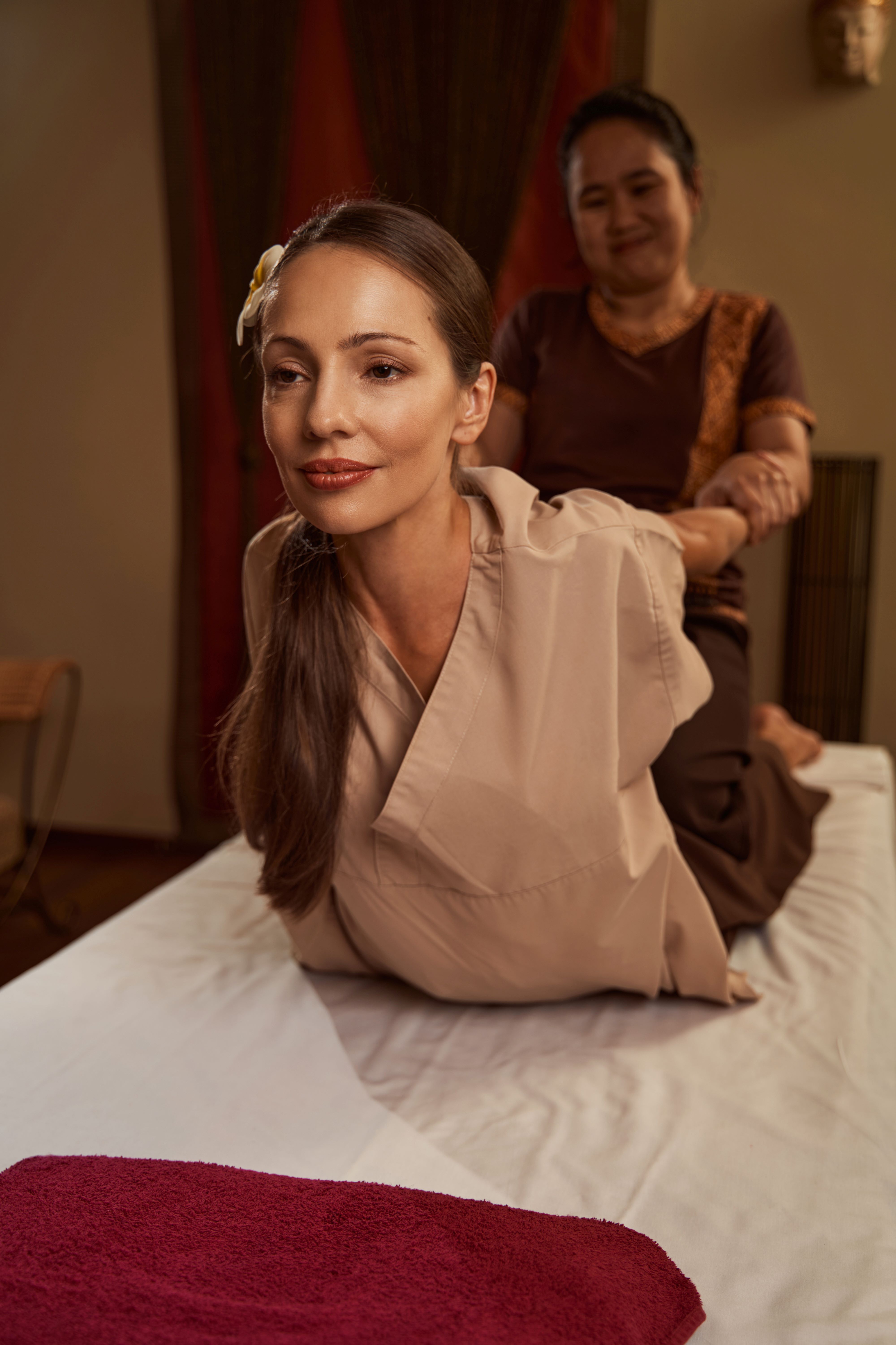 A woman laying on top of a bed being lifted by her arms and bent backwards, as part of a Thai massage.