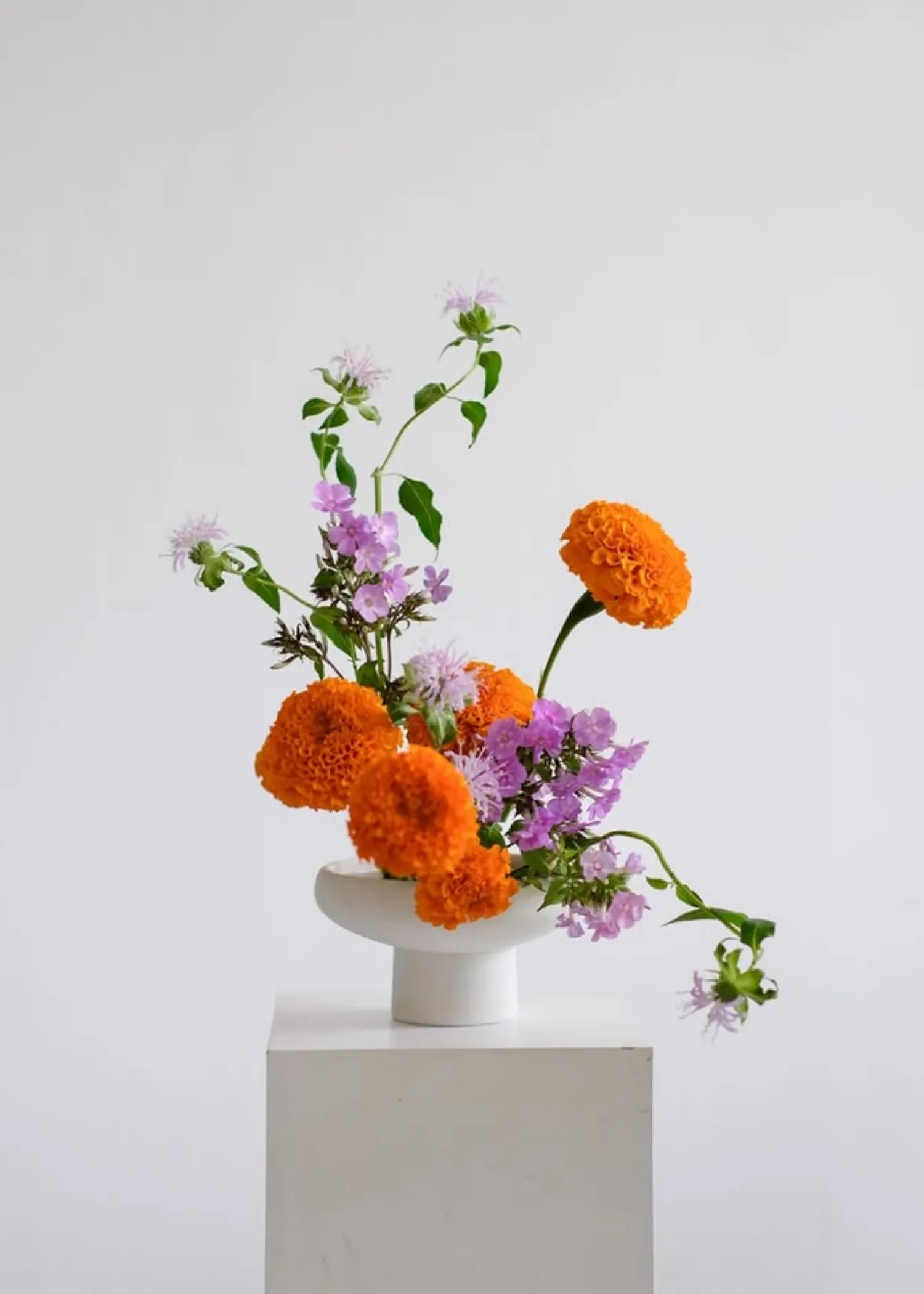 White pedestal holding a modern white vase with an arrangement of bright orange marigold flowers and delicate light purple flowers with green leaves.