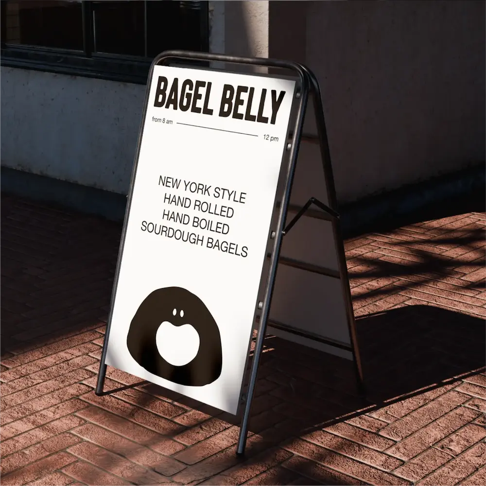 Outdoor sandwich board sign with text 'BAGEL BELLY' advertising New York style hand rolled, hand boiled sourdough bagels with a smiling bagel illustration at the bottom.
