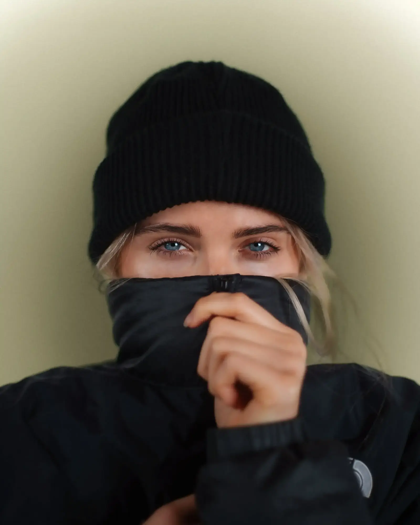 Person with blue eyes wearing a black knit hat and black jacket, covering lower face with their jacket collar.